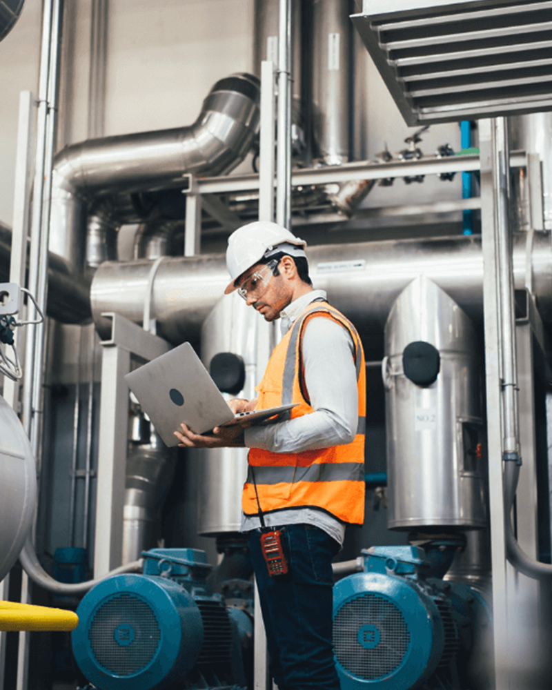Engineer wearing safety vest and hard hat using a laptop to monitor industrial piping and machinery in a factory setting
